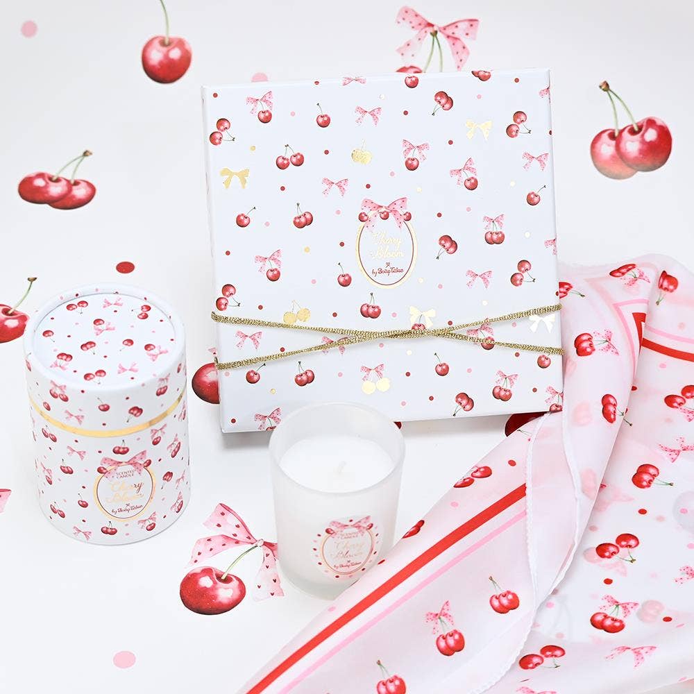 Cherry Bloom scented candle by SoapTales, featuring a frosted glass candle holder next to a decorative cylindrical gift tube illustrated with red cherries, pink bows, and gold accents.