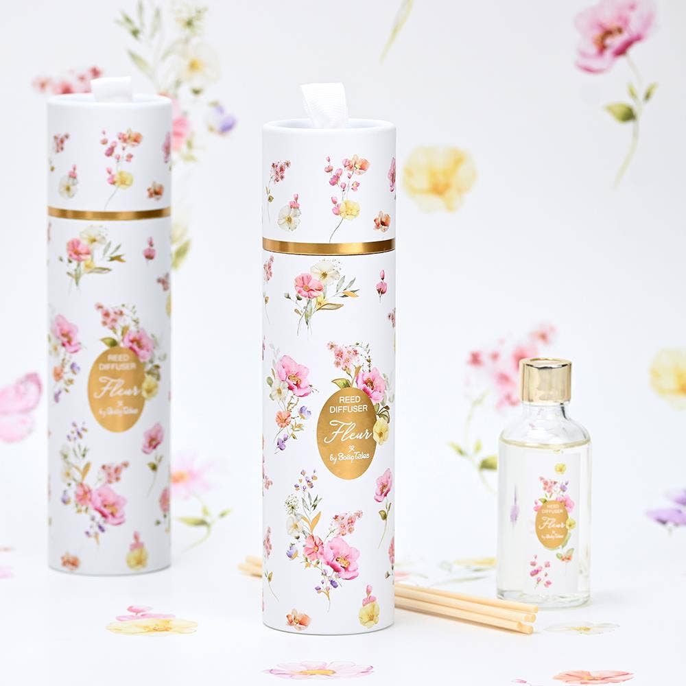 Fleur Pink Reed Diffuser by SoapTales, featuring a clear glass bottle with wooden reeds and a decorative floral cylindrical box in pink, yellow, and lilac hues.